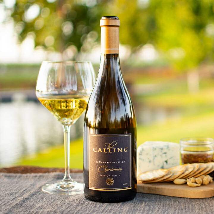 Bottle of one of The Calling's Chardonnays displayed on table, with crackers and cheese on its right and glass of wine on its left