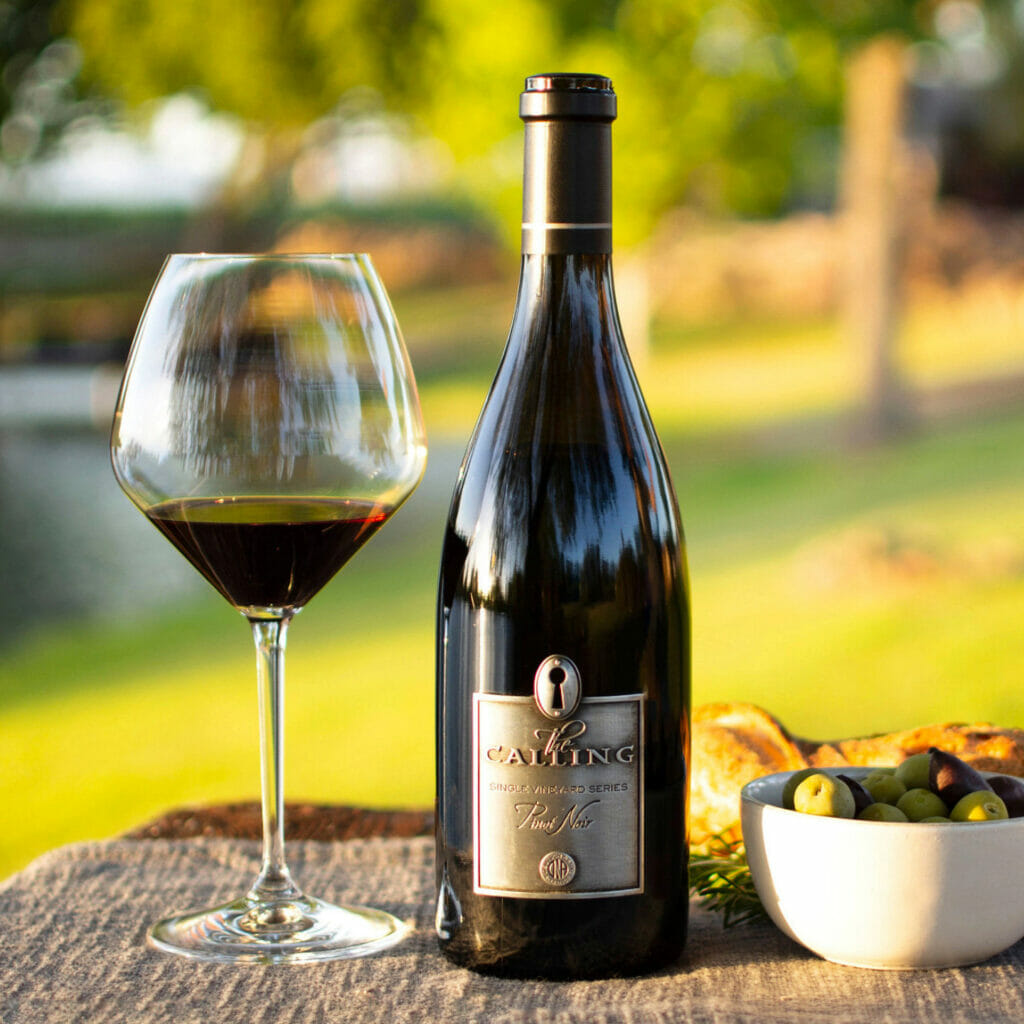 Bottle of Pinot Noir from the Calling displayed on table, with a glass of red wine and olives on both sides.